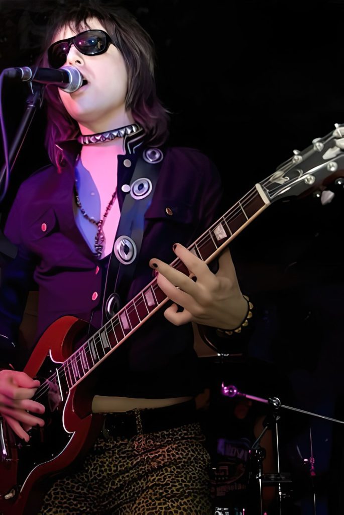 Sylvain Peter sur scène dans sa période glam rock, chantant et jouant de la guitare avec des lunettes de soleil et un pantalon léopard.